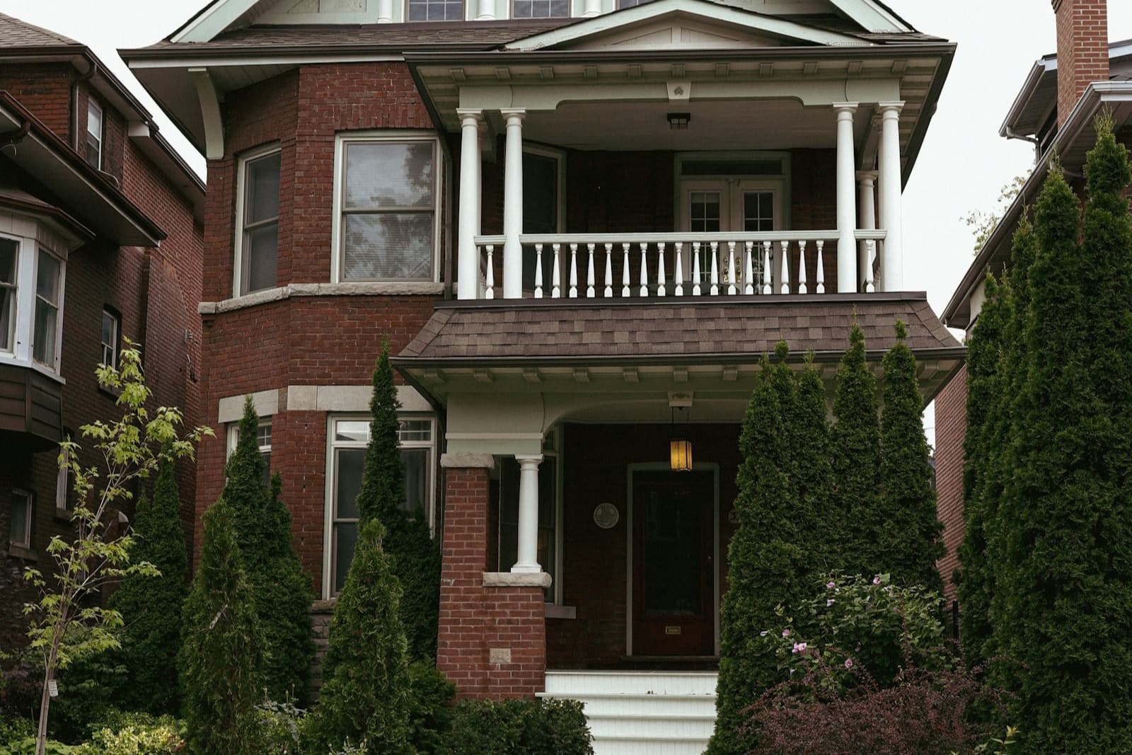 A red-brick two-storey Toronto home with a columned porch, white balcony railing, and manicured cedars out front